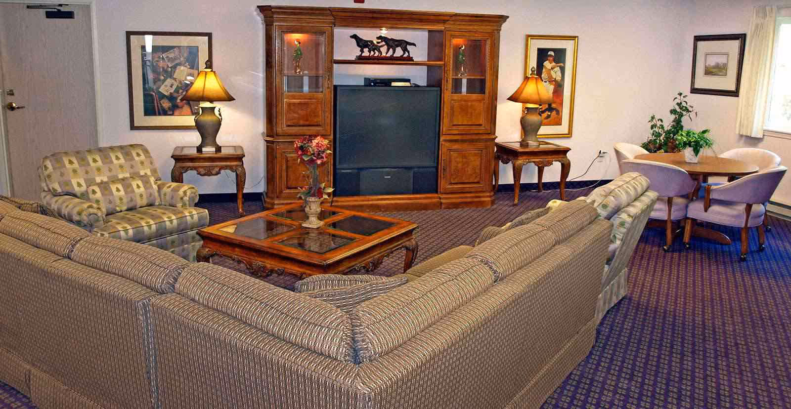 A cozy communal living room with sofas and armchairs arranged around a coffee table facing a wooden entertainment center with a TV.