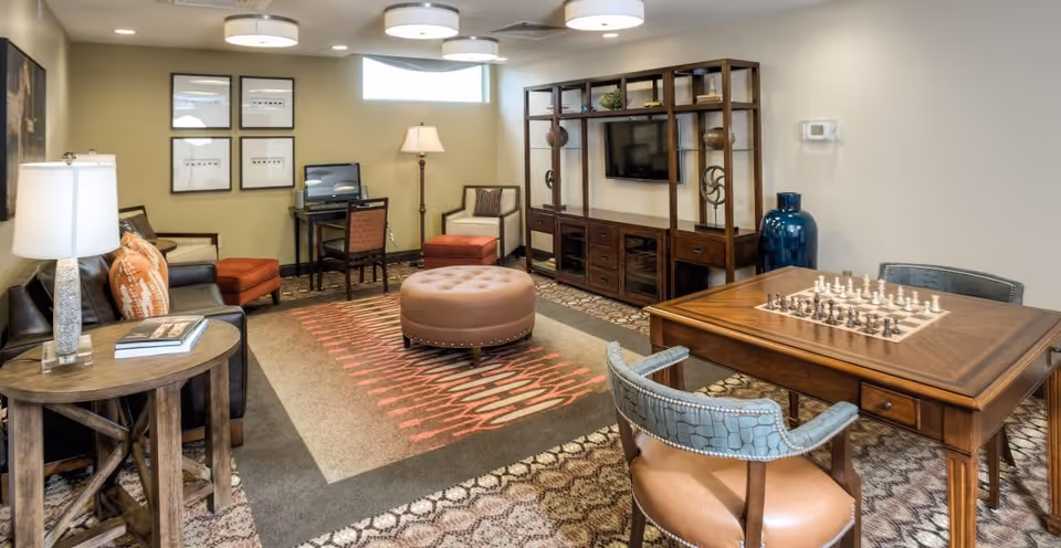Communal living room with sofa, chairs, ottoman, TV shelving unit and a chess table.