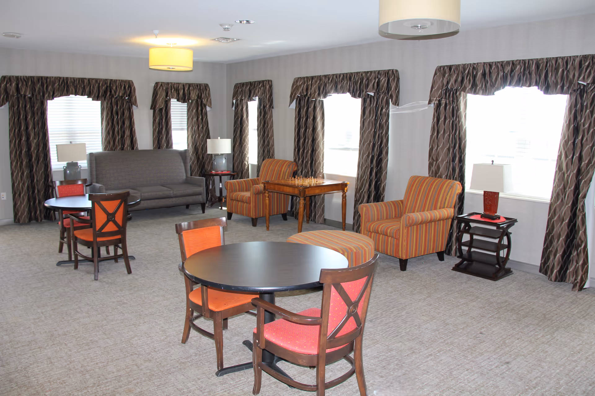 A spacious common area with multiple windows covered by patterned curtains. The room features a gray sofa, two striped armchairs with a matching ottoman, several round tables with wooden chairs that have red cushions, and side tables with lamps. The carpet is light-colored and the walls are neutral-toned.