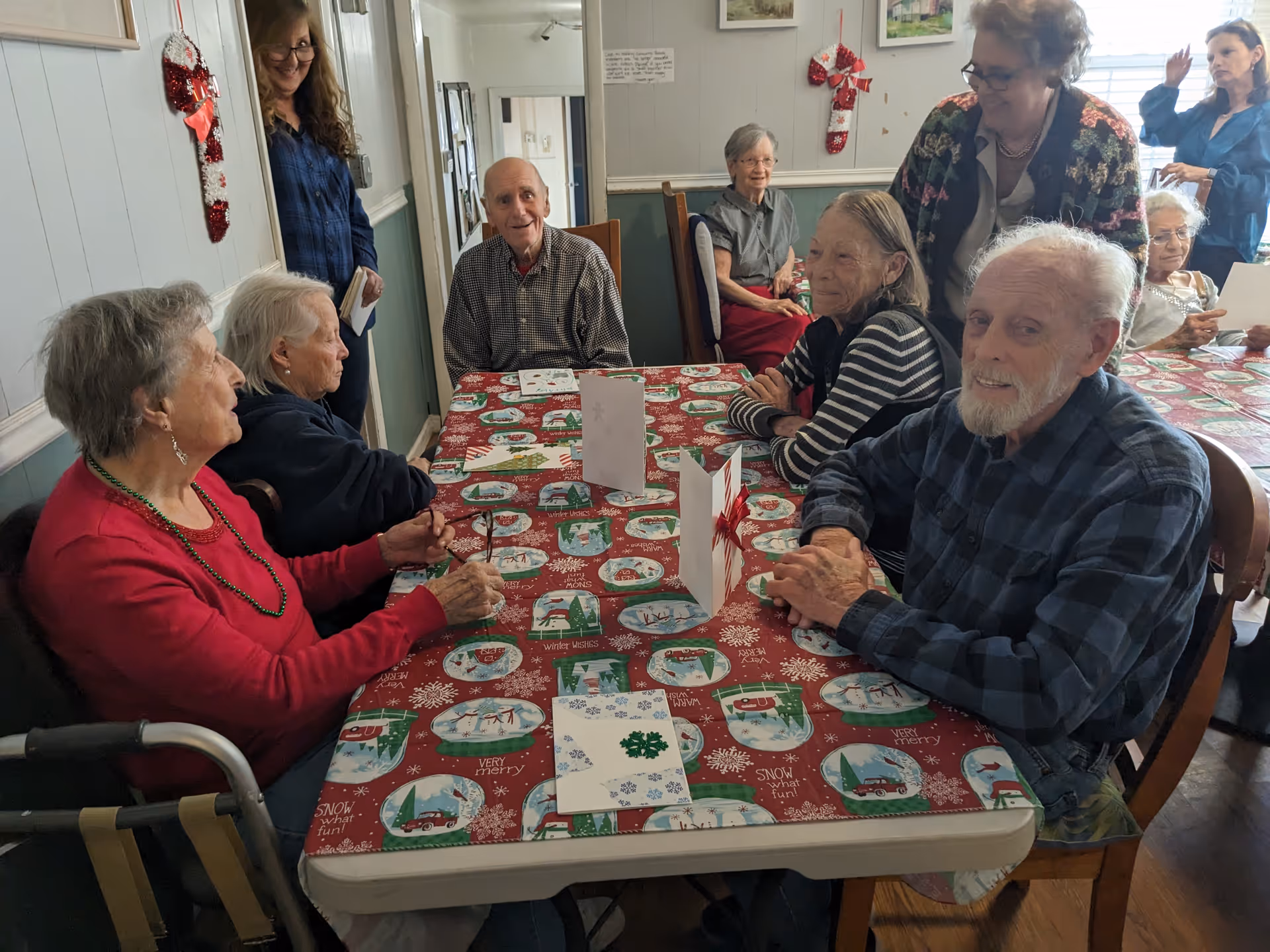A group of elderly people sitting around a table covered with a festive holiday tablecloth, engaging in conversation and smiling. Two women stand nearby, one holding a book. The room has light-colored walls decorated with holiday stockings and framed pictures.