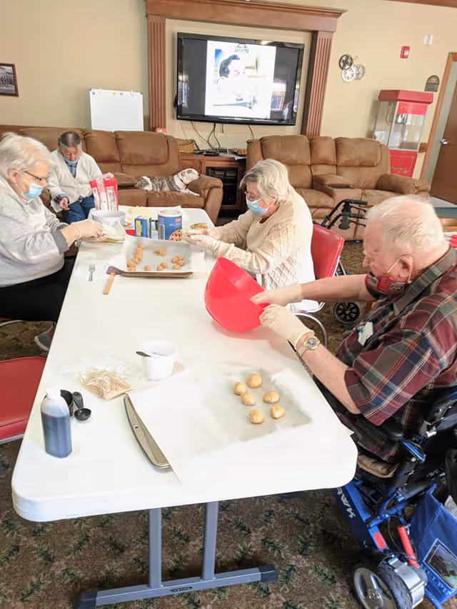 Four elderly individuals wearing masks and gloves are seated around a white table in a living room setting, preparing cookie dough balls on baking sheets. Behind them are brown couches, a television mounted on the wall, and a popcorn machine. One person is in a wheelchair.