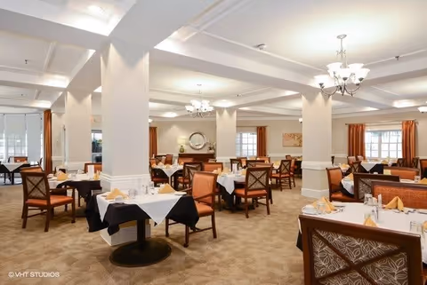 A spacious dining room with multiple tables covered with white and black tablecloths, set with napkins, glasses, and silverware. The room features beige carpet, white walls with columns, large windows with orange curtains, and elegant chandeliers hanging from the ceiling.