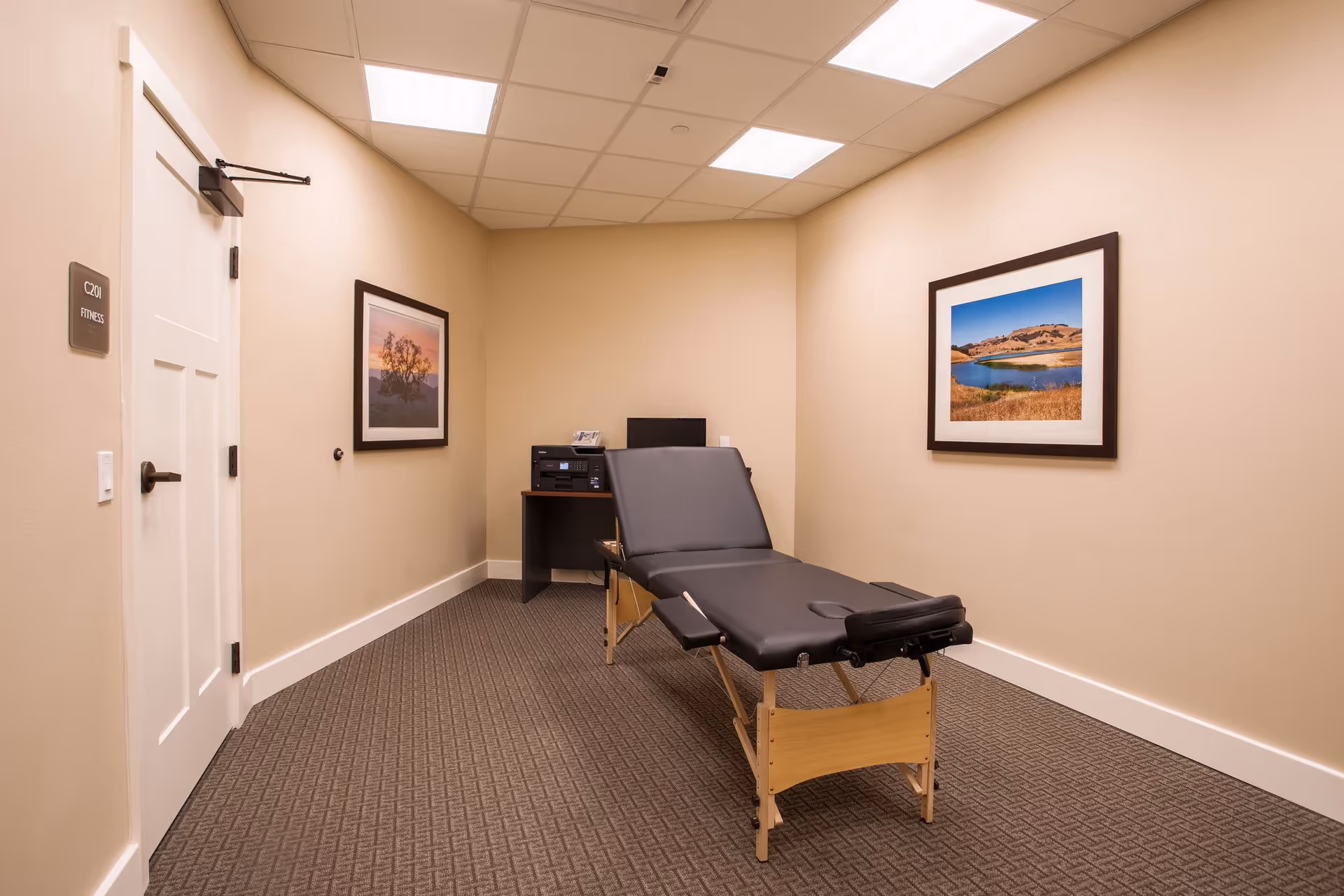 A small room with beige walls and carpeted floor featuring a black massage or therapy table in the center. On the left wall, there is a white door labeled 'C201 FITNESS'. The room has two framed landscape photographs on the walls and a small desk with a printer and a computer monitor in the back corner.