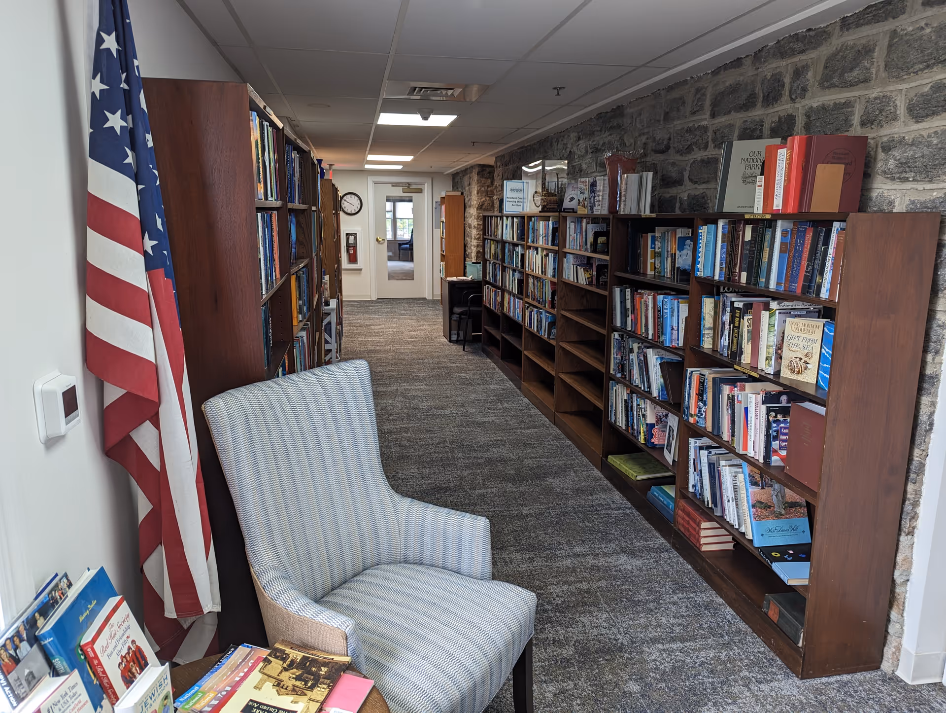 Interior hallway of a senior living facility featuring bookshelves filled with books along a stone wall, a comfortable upholstered chair, an American flag, and a carpeted floor leading to a door at the end of the corridor.