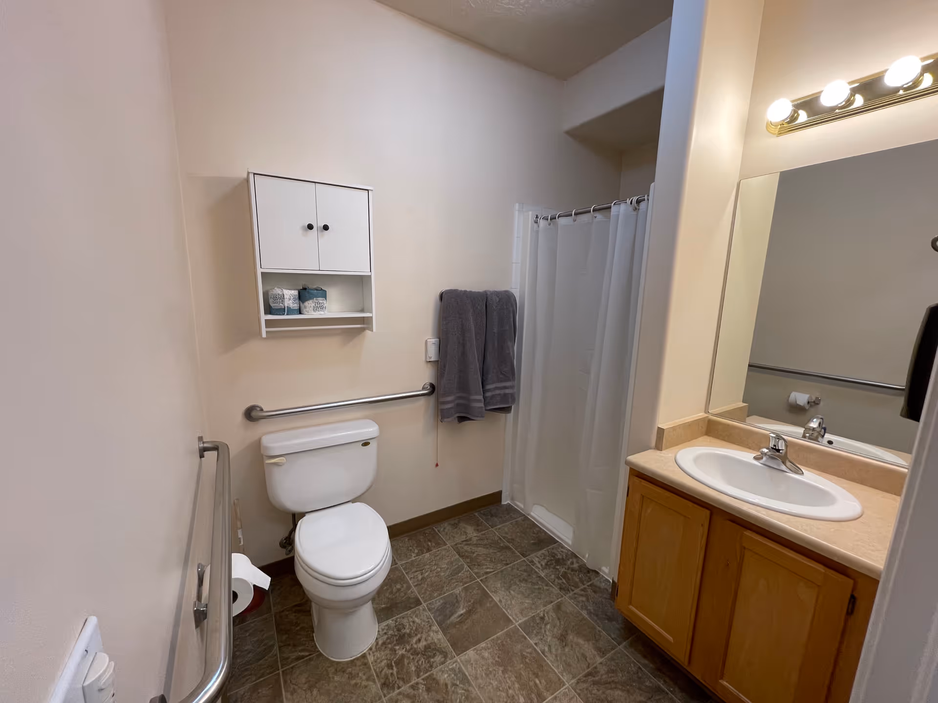 A clean and accessible bathroom with a white toilet, grab bars on the walls, a small white cabinet mounted above the toilet, a shower with a white curtain, a sink with a wooden cabinet below, and a large mirror with lights above it.