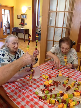 Two elderly women sitting at a table covered with a red and white checkered tablecloth, making fruit skewers with various fruits like grapes, pineapple, and melon. One woman is holding a skewer upright, while the other is reaching for fruit. The room has wooden doors and yellow walls with framed pictures and a small table with a flower vase in the background.