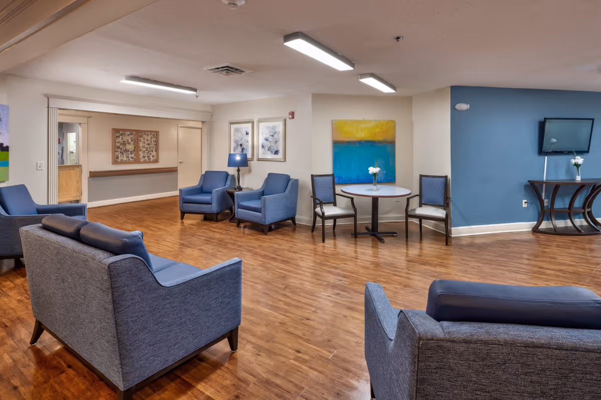 A spacious common area with wooden flooring, featuring several blue upholstered armchairs and sofas arranged for seating. There is a small round table with two chairs and a vase with flowers, a blue wall with a mounted TV, and colorful artwork on the walls. The room is well-lit with ceiling lights.
