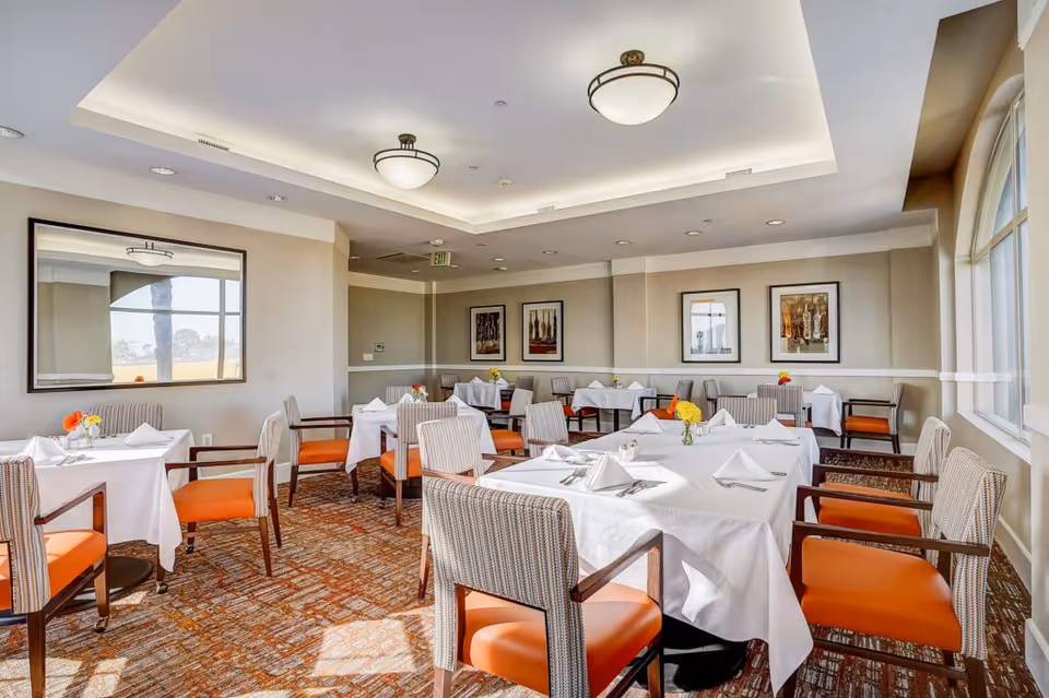 A bright dining room with several tables covered in white tablecloths, each set with napkins, silverware, and small flower arrangements. The chairs have orange cushions and patterned backs. The room has beige walls adorned with framed artwork and a large window letting in natural light.