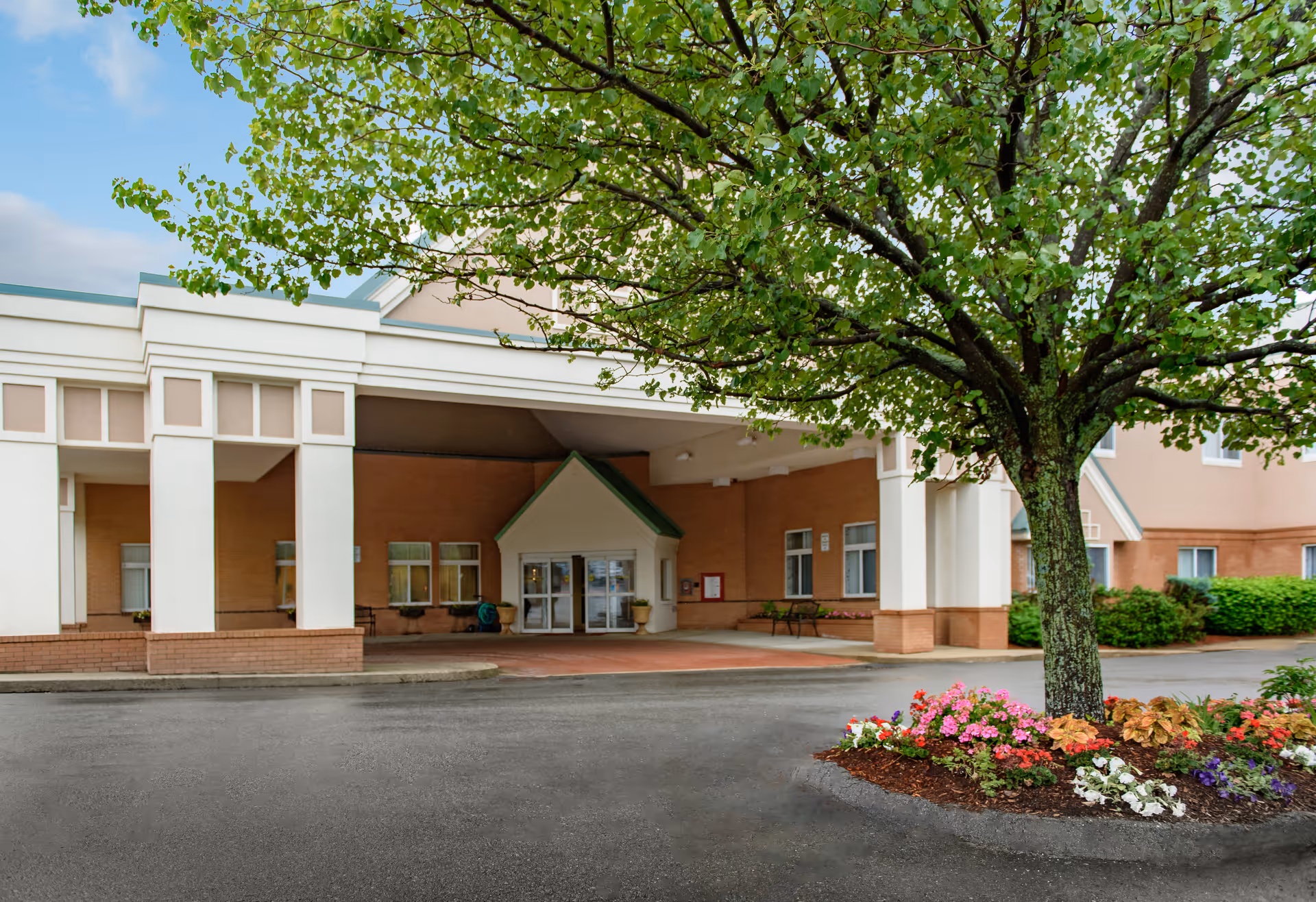 Entrance of a senior living facility with a covered drop-off area, a large tree with green leaves, and a flower bed with colorful flowers in front. The building has beige and white exterior walls with multiple windows.