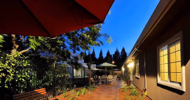 Outdoor patio area at dusk with a paved walkway, patio tables and chairs under umbrellas, a bench, plants along the path, and a building with lit windows on the right side.