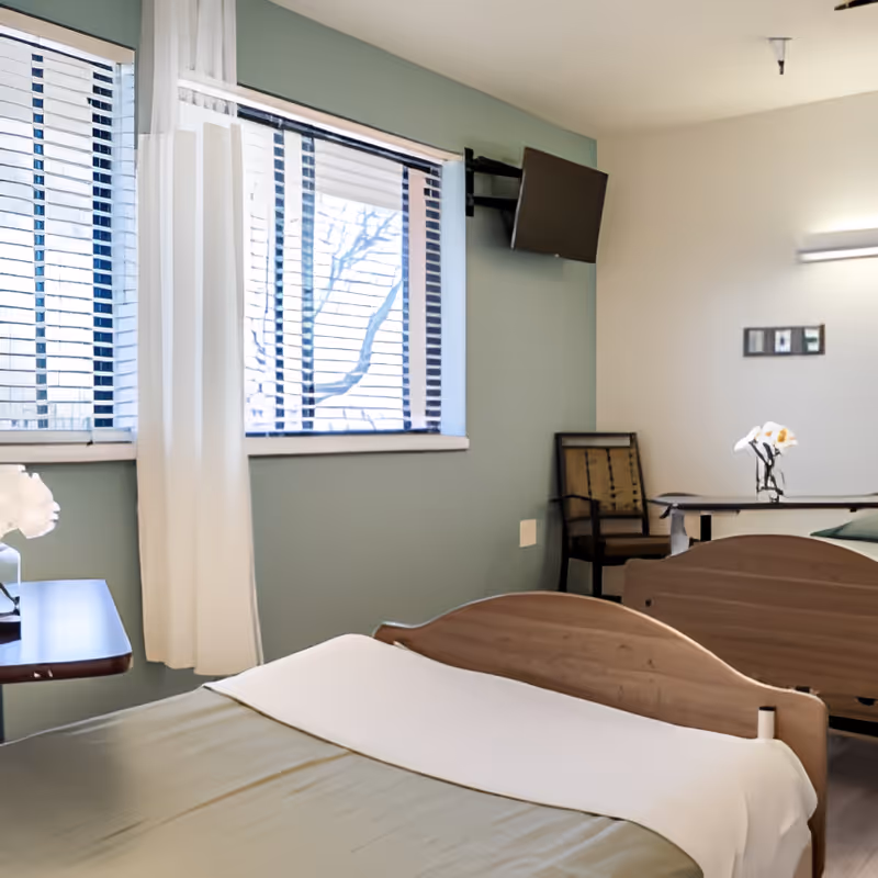 A bright shared bedroom with two beds, a window with blinds, a wall-mounted TV, a chair, and a bedside table with flowers.