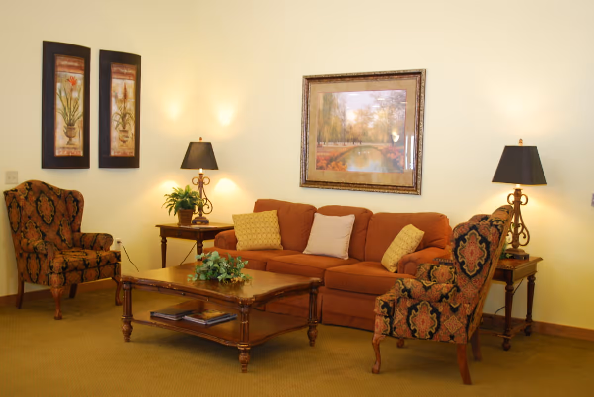 A cozy living room with a rust-colored sofa adorned with three pillows, two patterned armchairs, a wooden coffee table with a plant centerpiece, two side tables each with a lamp, and framed artwork on the walls.