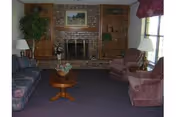 A living room with a brick fireplace centered on the back wall, flanked by built-in wooden shelves. The room has a purple carpet, a wooden coffee table with a flower arrangement, two purple upholstered armchairs, a patterned sofa, two white lamps, a large potted plant, and a window with a valance on the right side.