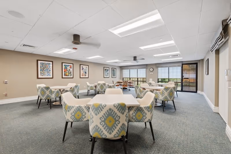 A spacious dining room with multiple square tables surrounded by patterned upholstered chairs. The room has a carpeted floor, beige walls adorned with framed artwork, and large windows with blinds allowing natural light. Ceiling fans and recessed lighting are installed on the white ceiling. A clock is mounted on the far wall near a glass door leading outside.