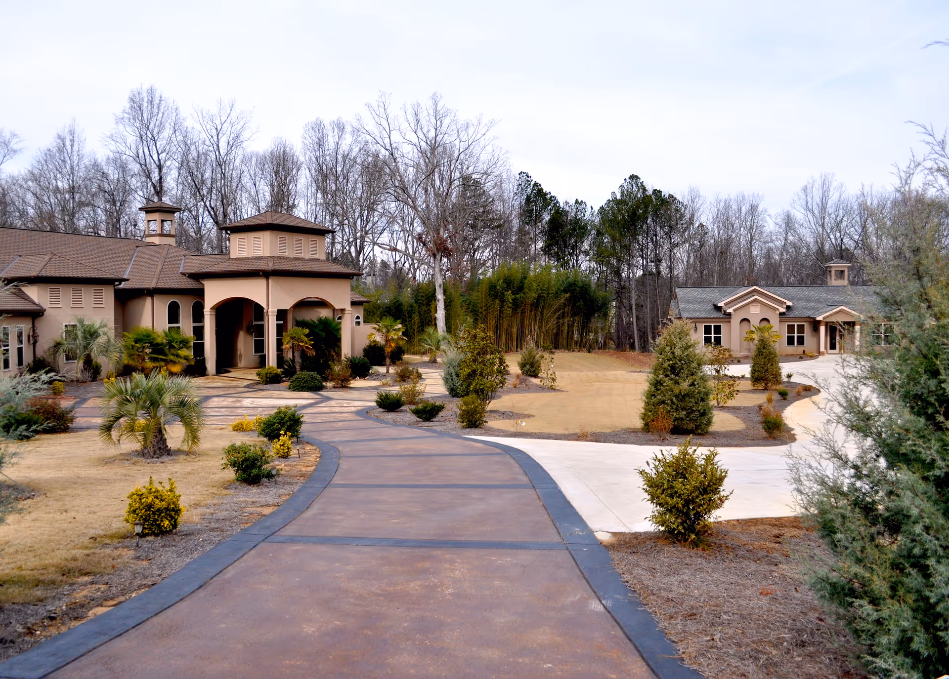 View of the front exterior of a senior living facility with a curved driveway leading to two beige buildings surrounded by landscaped bushes and trees, with leafless trees in the background under a cloudy sky.