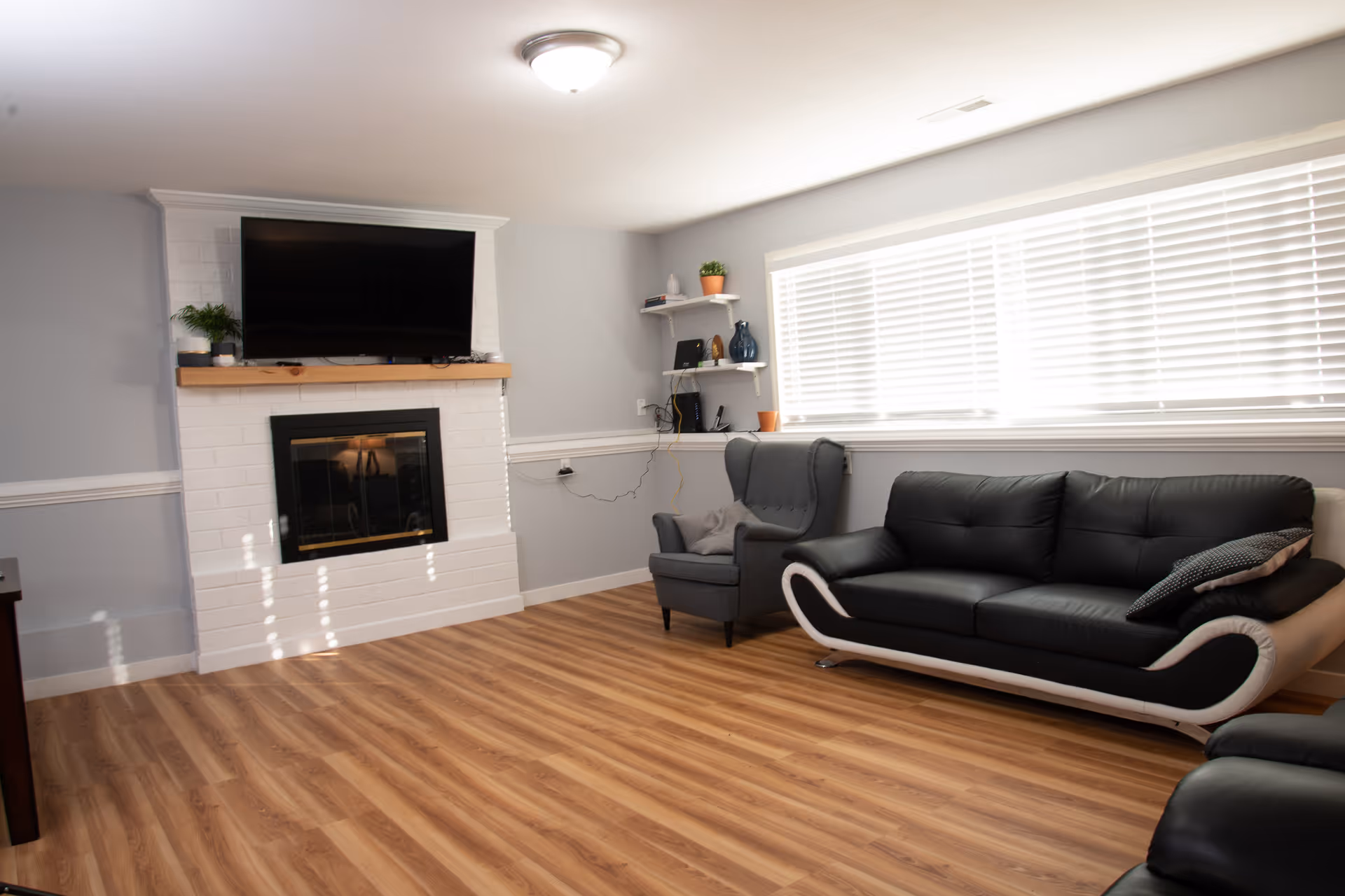 Bright modern living room with a white brick fireplace and mounted TV, black leather sofa and armchair, and a large window with blinds.