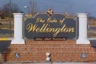 A large sign for The Oaks of Wellington, a senior adult community, mounted on a brick base with two white columns on either side, located outdoors with a clear sky background.