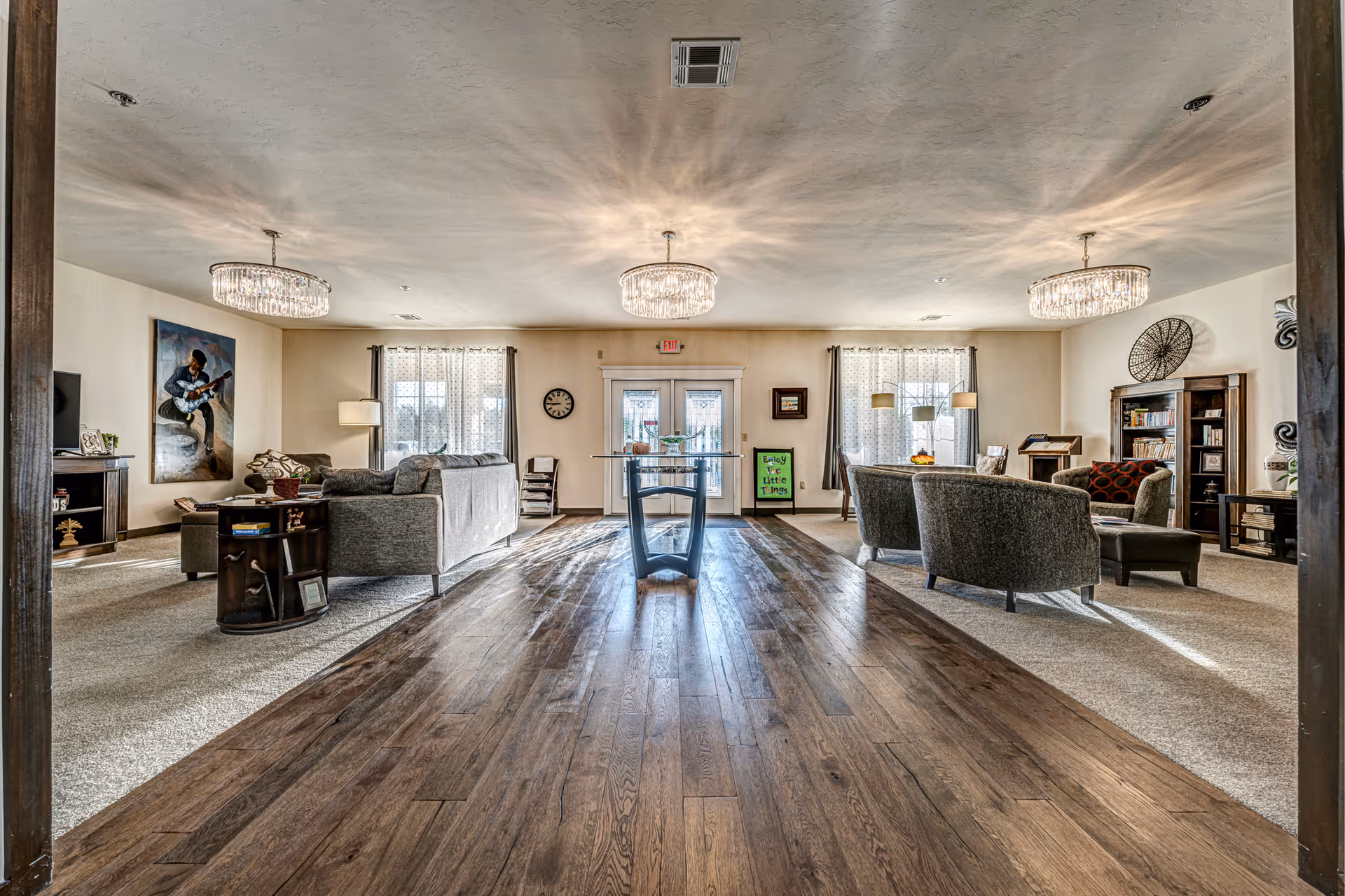 Spacious senior living common area with wooden flooring in the center and carpeted seating areas on both sides. The room features modern chandeliers, large windows with sheer curtains, comfortable sofas and chairs, bookshelves, and decorative artwork on the walls. A glass table is positioned in the middle of the wooden floor section.