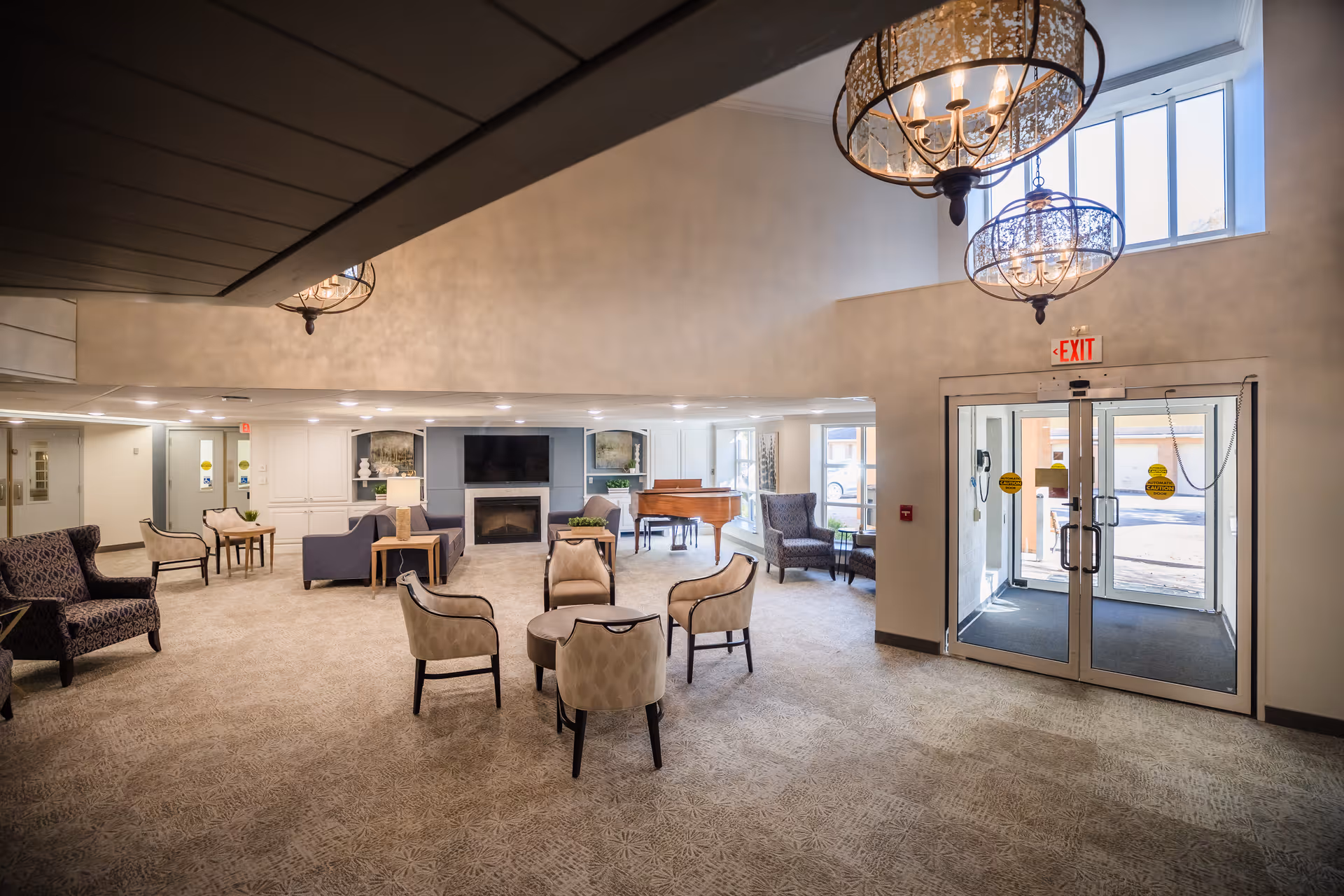 Spacious and well-lit common area in a senior living facility featuring multiple seating arrangements with armchairs and small tables, a fireplace with a TV mounted above it, a grand piano near large windows, and glass double doors leading outside. The room has high ceilings with decorative hanging light fixtures and carpeted flooring.