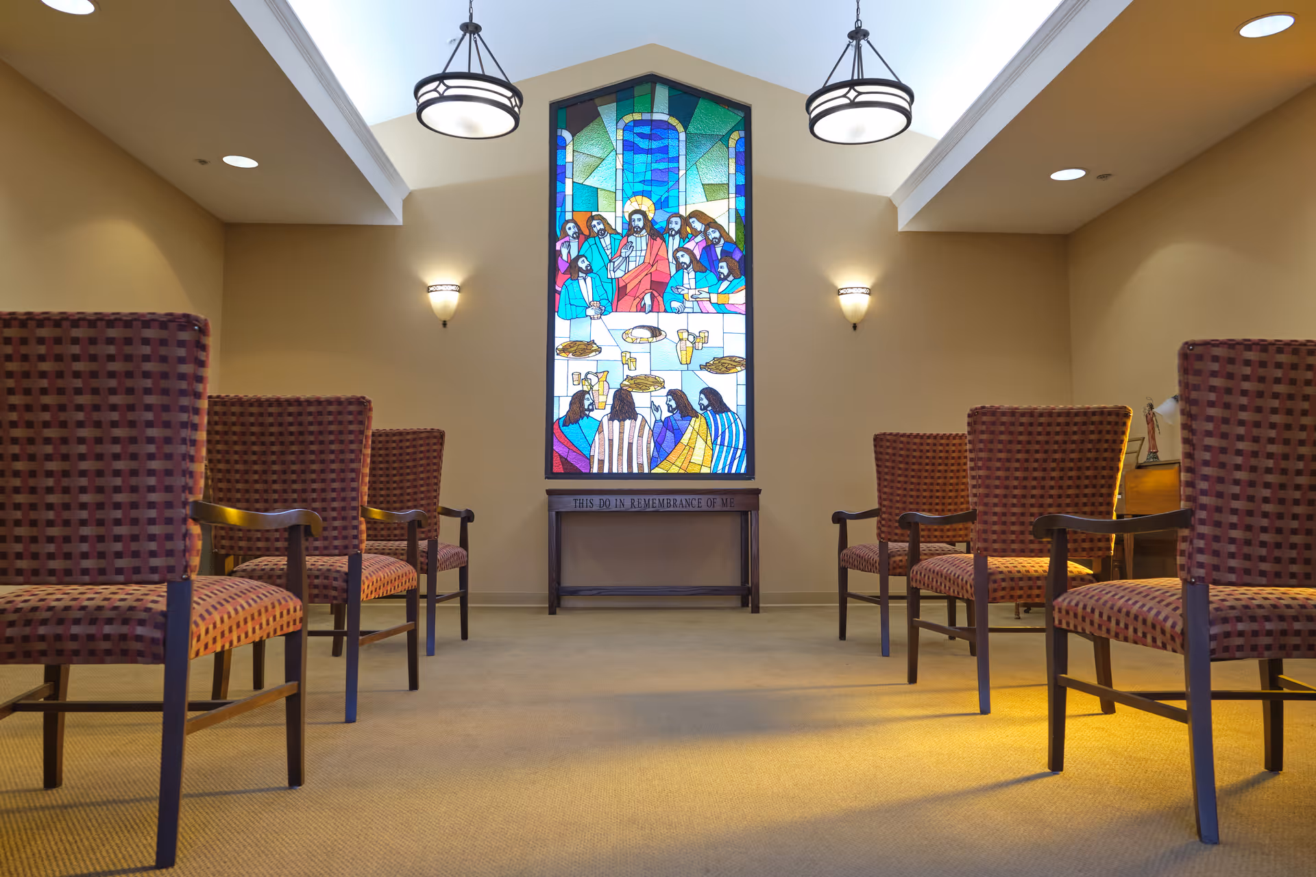 Interior room with rows of patterned chairs facing a stained glass window depicting a religious scene. The room has beige walls, carpeted floor, and ceiling lights. Below the stained glass window is a small wooden table with the inscription 'THIS DO IN REMEMBRANCE OF ME'.