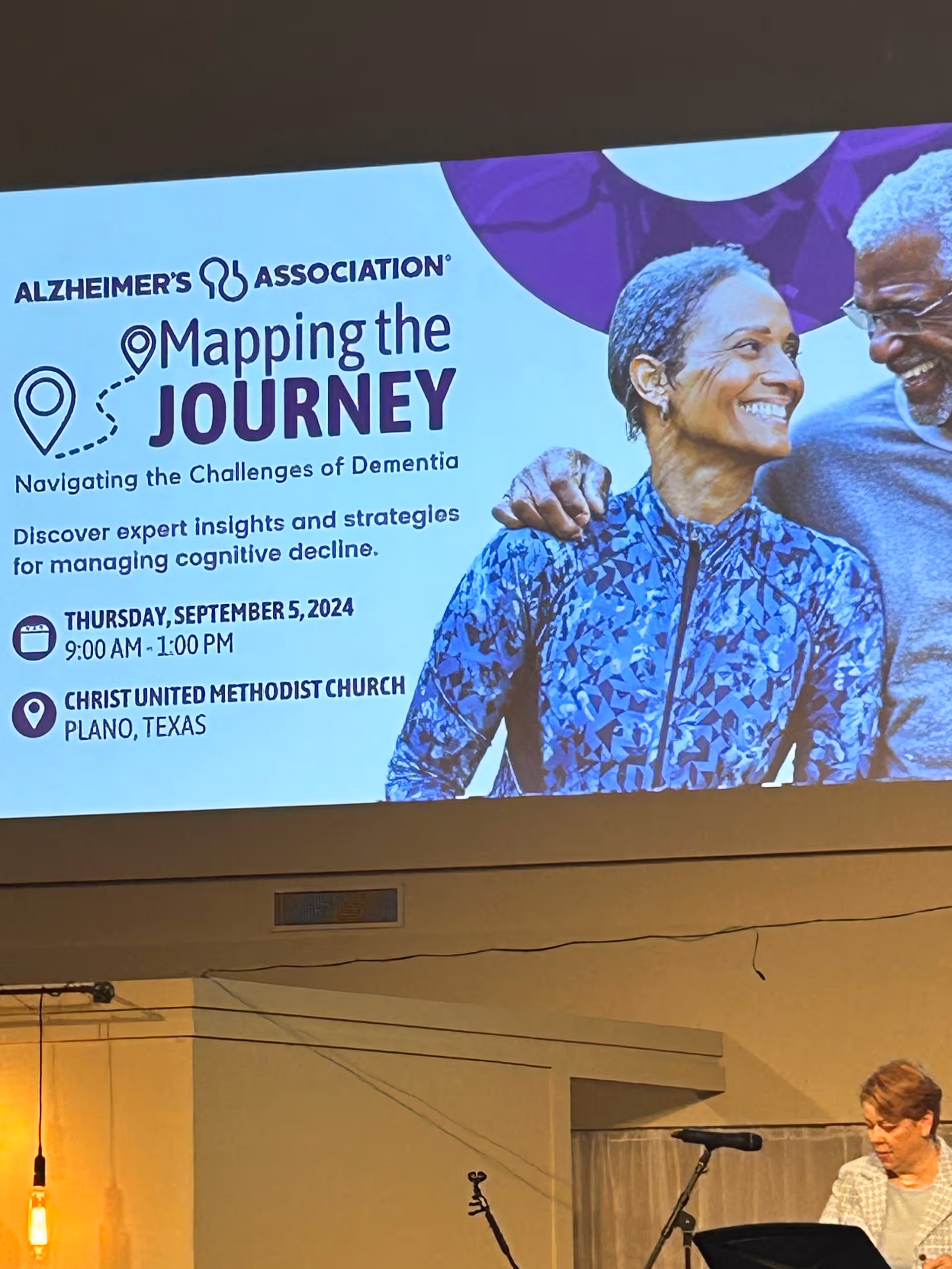 A presentation slide from the Alzheimer's Association titled 'Mapping the Journey: Navigating the Challenges of Dementia' with details about an event on Thursday, September 5, 2024, from 9:00 AM to 1:00 PM at Christ United Methodist Church in Plano, Texas. The slide features a smiling older couple on the right side. Below the slide, a woman stands at a podium with a microphone in a room with beige walls and hanging lights.