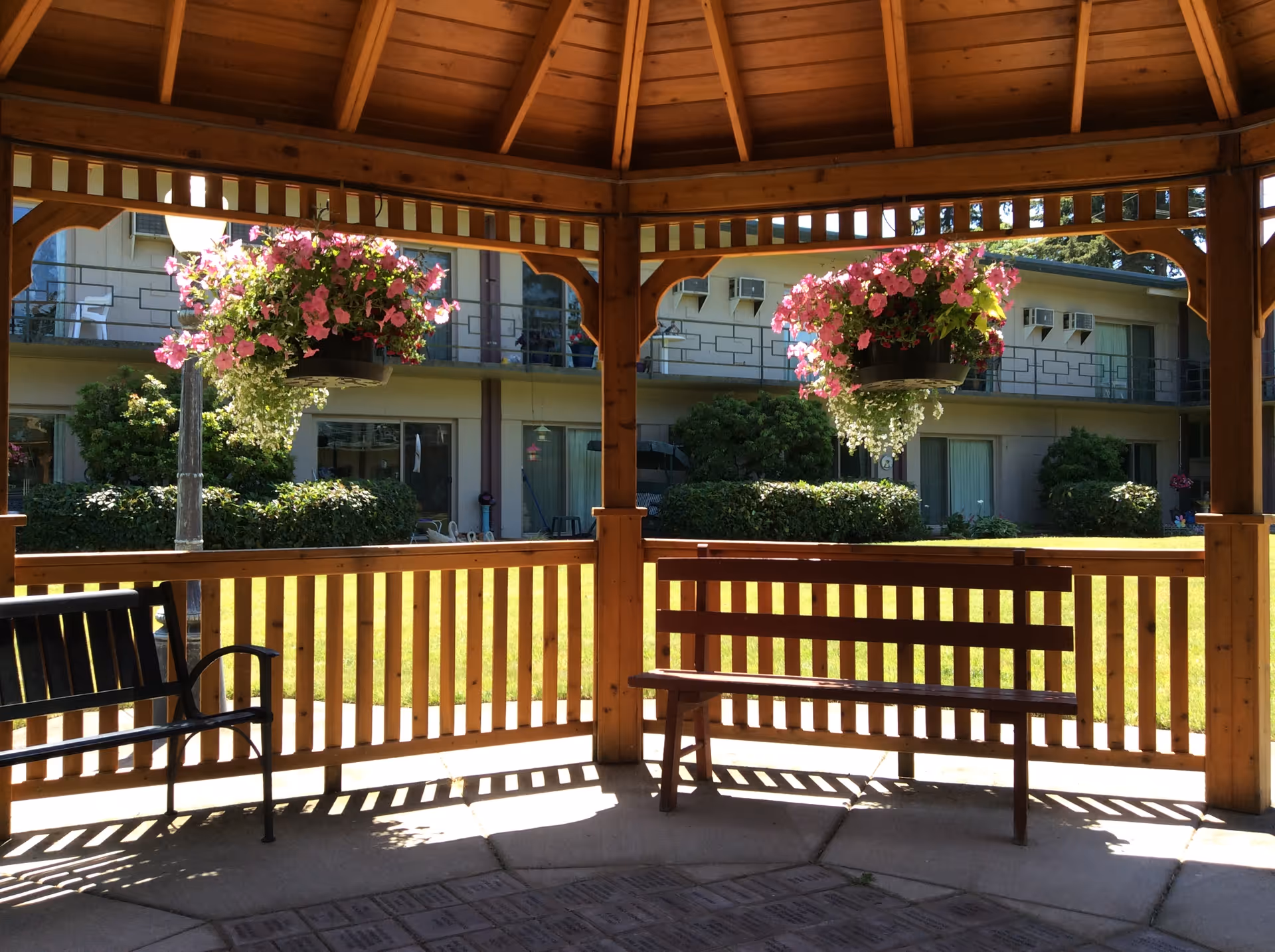 View from inside a wooden gazebo with a bench and a metal chair, overlooking a grassy courtyard with hanging flower baskets and a two-story building with balconies and windows in the background.