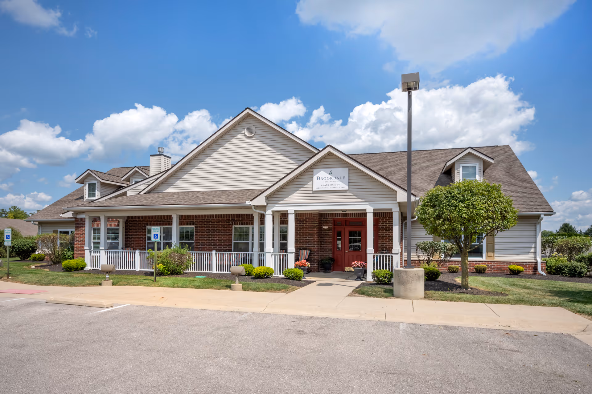 Front exterior of the Brookdale Greenville senior living building with a covered porch entrance, red doors, and landscaped grounds under a blue sky.