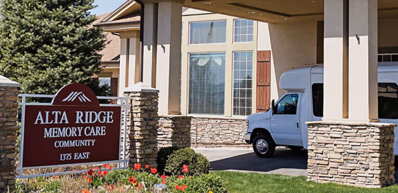 Exterior view of Alta Ridge Memory Care Community showing a covered driveway with a white van parked under it. The building has stone pillars and large windows with wooden shutters. In front, there is a sign with the facility name and address surrounded by flowers and greenery.