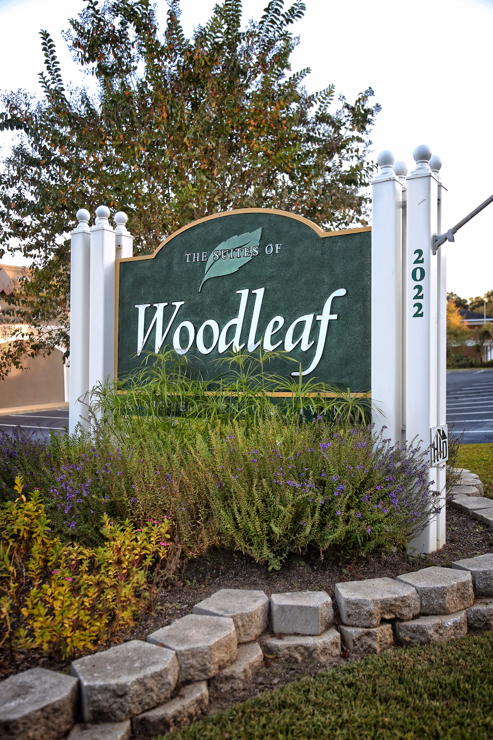A green and white outdoor sign for The Suites of Woodleaf, surrounded by plants and flowers with a tree in the background. The sign is mounted on white posts with the number 2022 on one post.