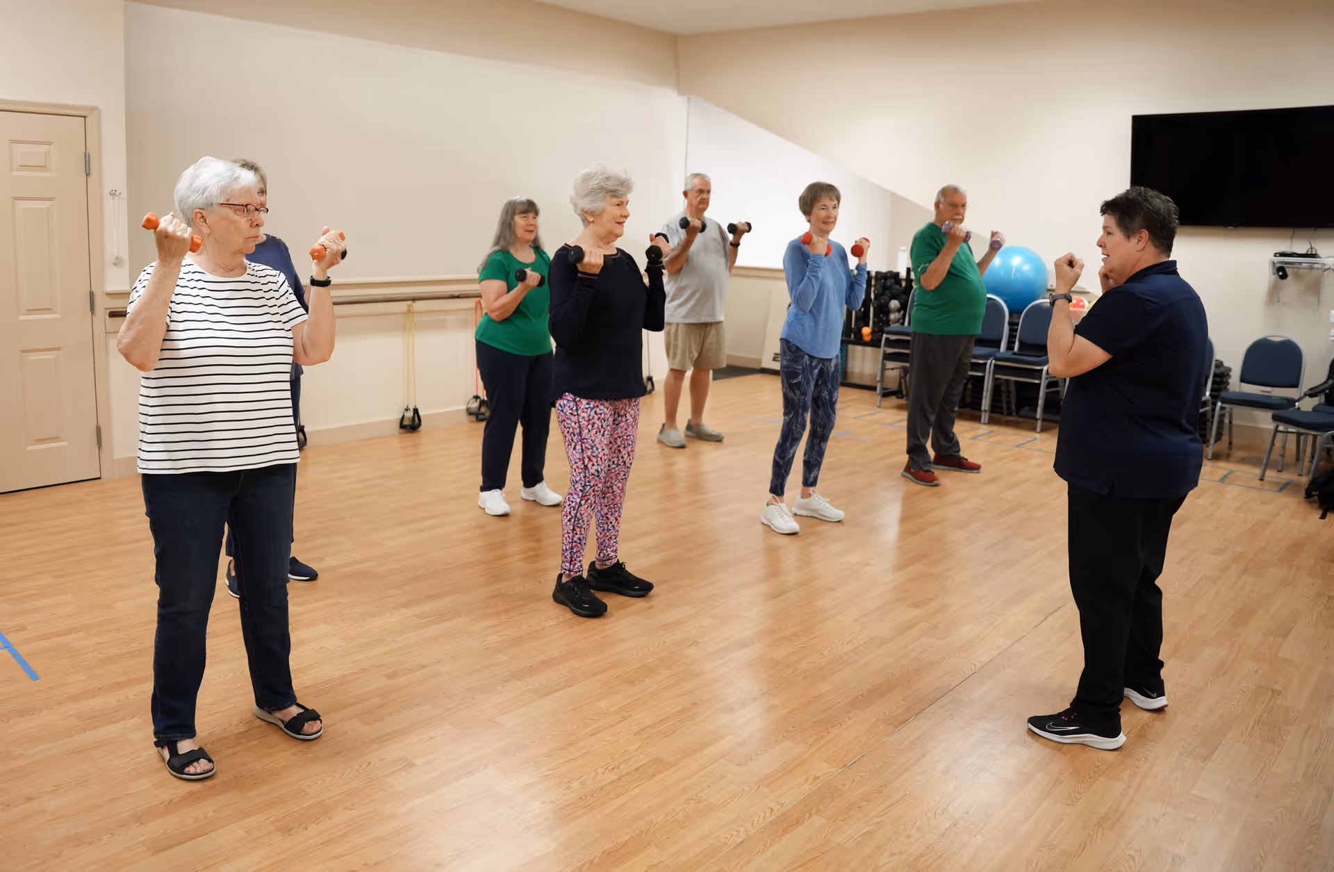A group of elderly individuals participating in a light exercise class indoors, each holding small dumbbells while following the instructor's guidance in a spacious room with wooden flooring and chairs along the walls.