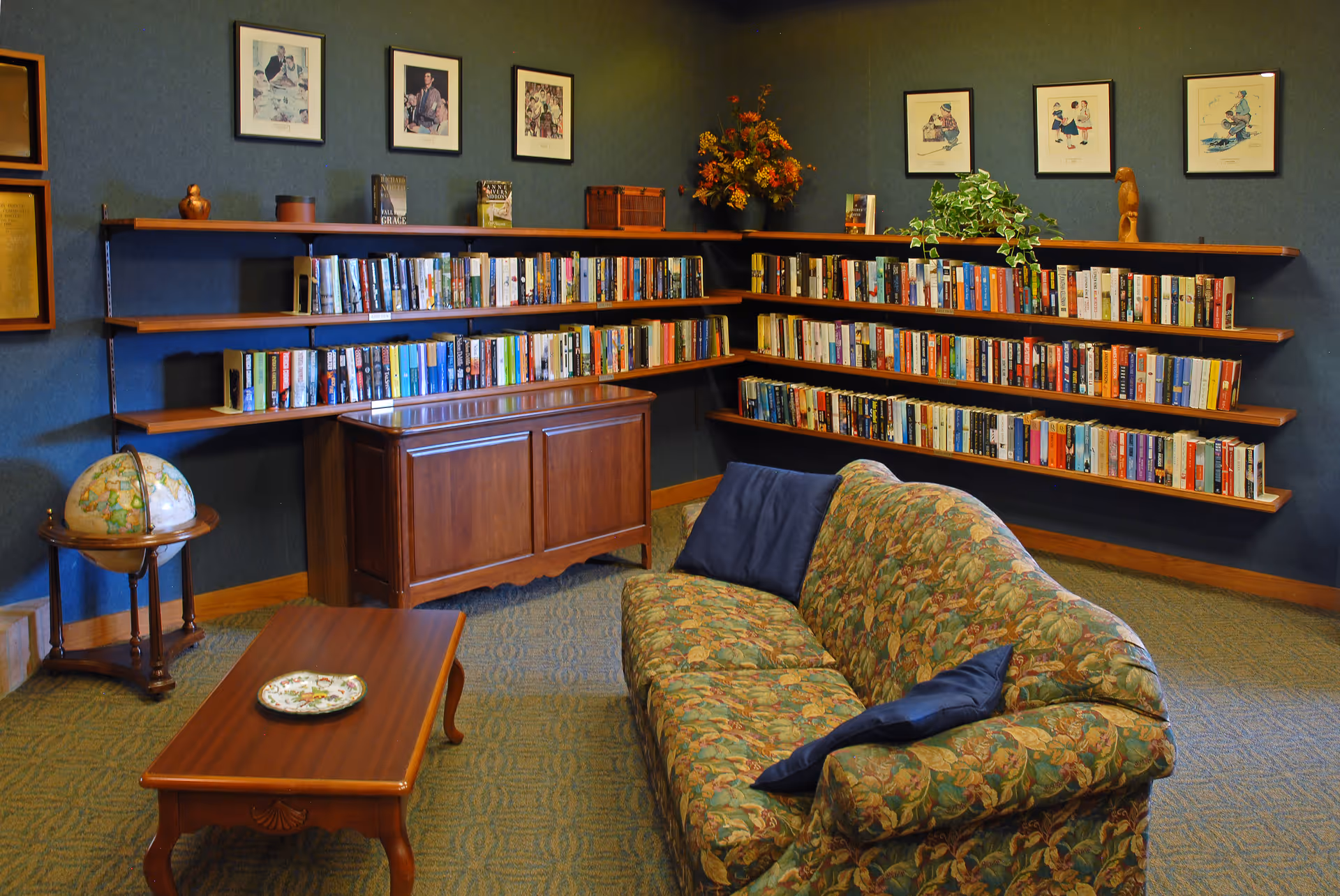 Cozy reading room with a floral patterned sofa adorned with two dark blue pillows, a wooden coffee table with a decorative plate, and corner wall-mounted bookshelves filled with books. The room has dark green walls decorated with framed pictures and a globe on a wooden stand in the corner.
