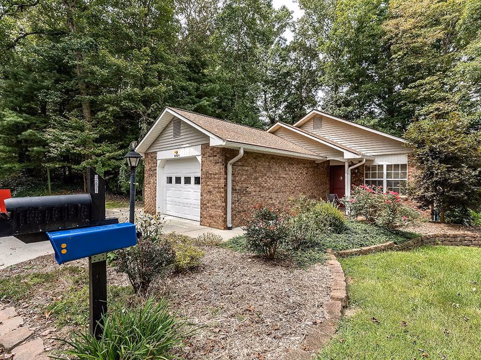 A single-story brick house with a white garage door and a red front door, surrounded by greenery and bushes. There is a blue mailbox and a black mailbox on a post near the driveway, and a lamppost next to the driveway. Trees and a lawn are visible around the house.