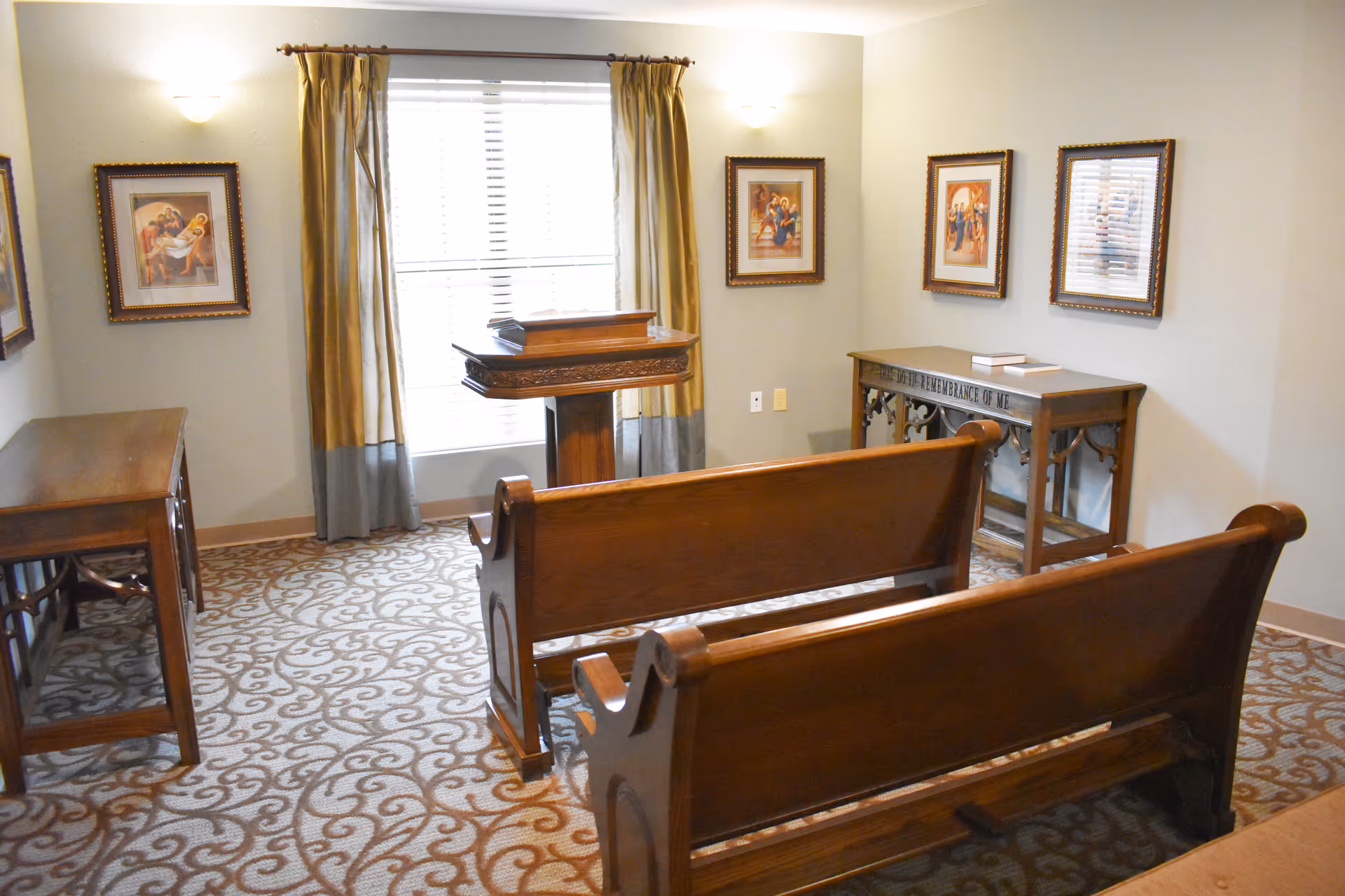 Small chapel-style interior with wooden pews facing a lectern by a window, framed religious artwork, and patterned carpet.