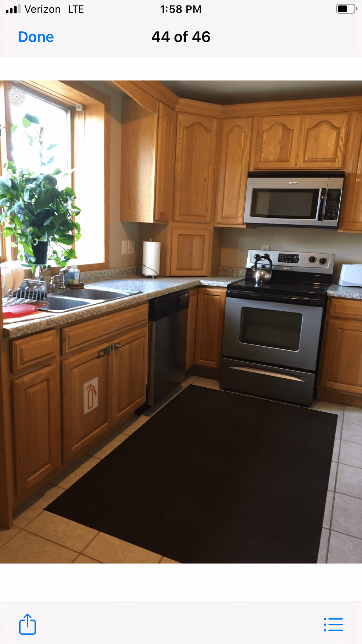 A kitchen with wooden cabinets, a stainless steel oven and microwave, a dishwasher, a double sink under a window with a green leafy plant on the windowsill, and a black floor mat on the tiled floor.
