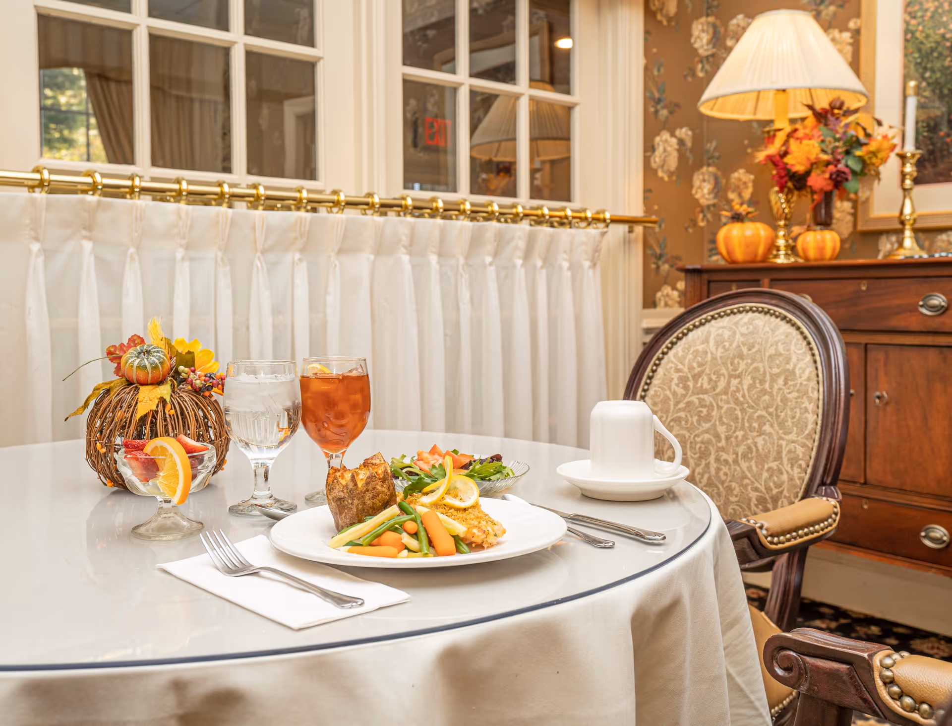 A round dining table set with a plate of food including vegetables and a baked potato, a glass of iced tea with lemon, a glass of water, a cup and saucer, and a small dessert dish with fruit garnished with an orange slice. The table is covered with a white tablecloth and is next to an upholstered chair. In the background, there is a wooden sideboard with a lamp, decorative pumpkins, and a floral arrangement, along with floral wallpaper and windows with white curtains.