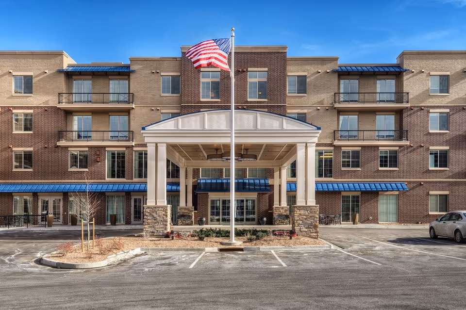 Front exterior view of a multi-story assisted living facility with a covered entrance, an American flag on a flagpole, balconies, and a parking area in front.