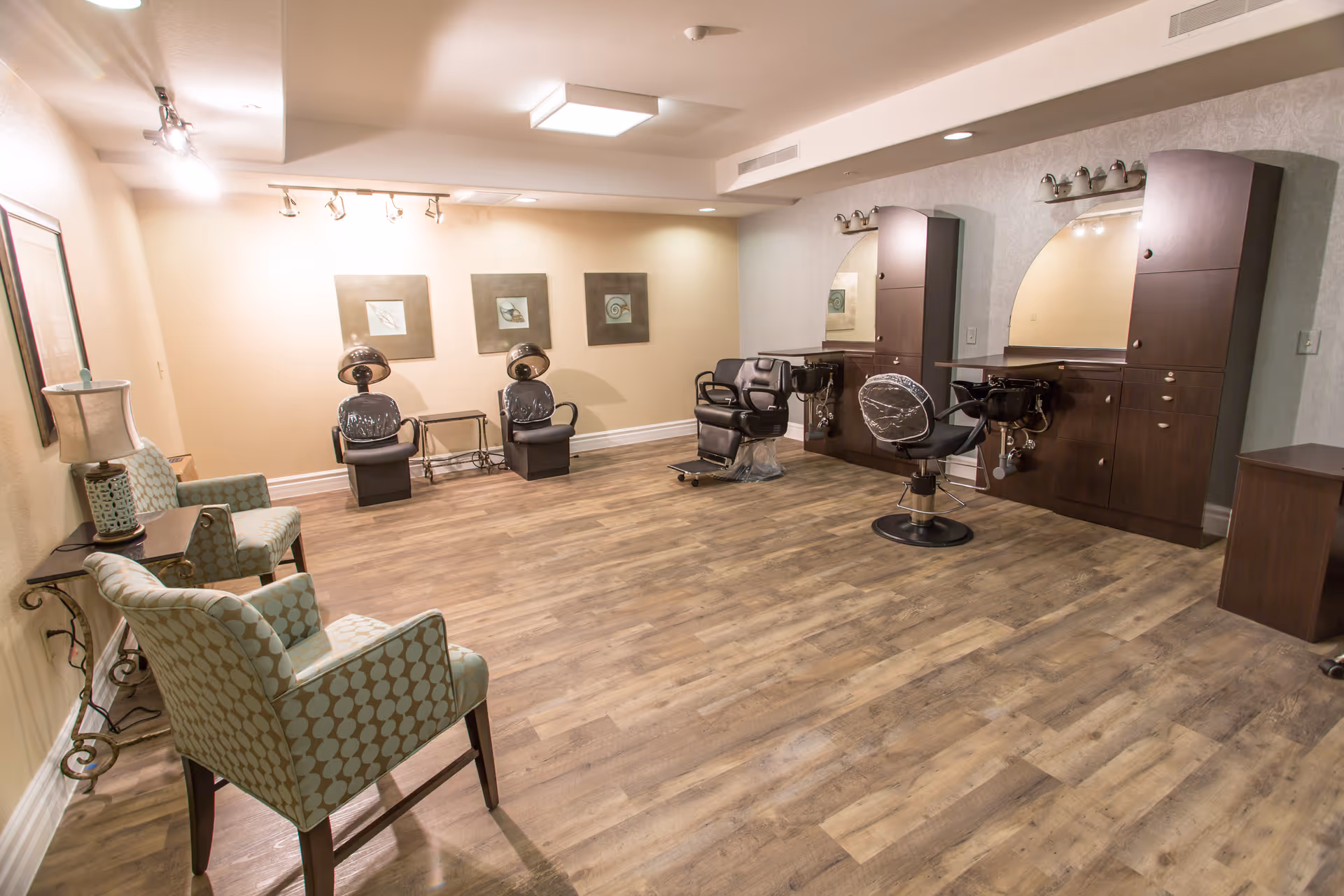 Empty hair salon area with styling chairs, hooded dryers, wash basins and lounge chairs on wood flooring.