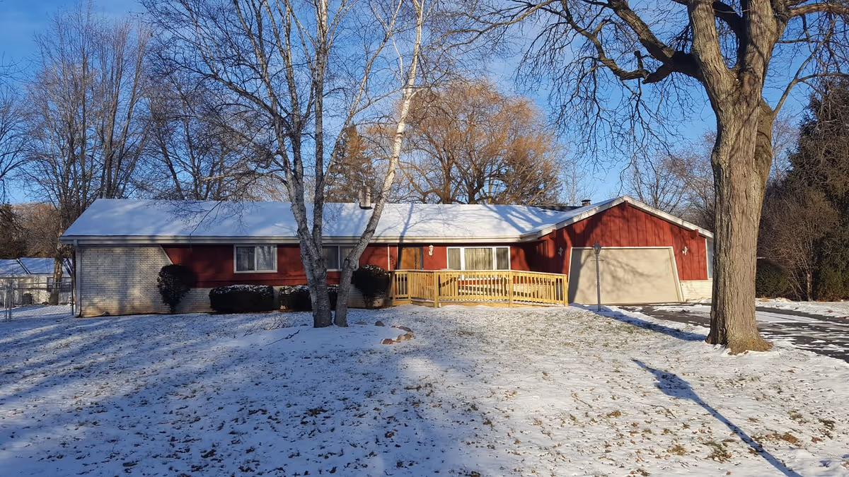 Single-story building with red siding and a white roof covered in a light layer of snow. There are leafless trees in the front yard and a wooden ramp leading to the entrance. The sky is clear and blue.