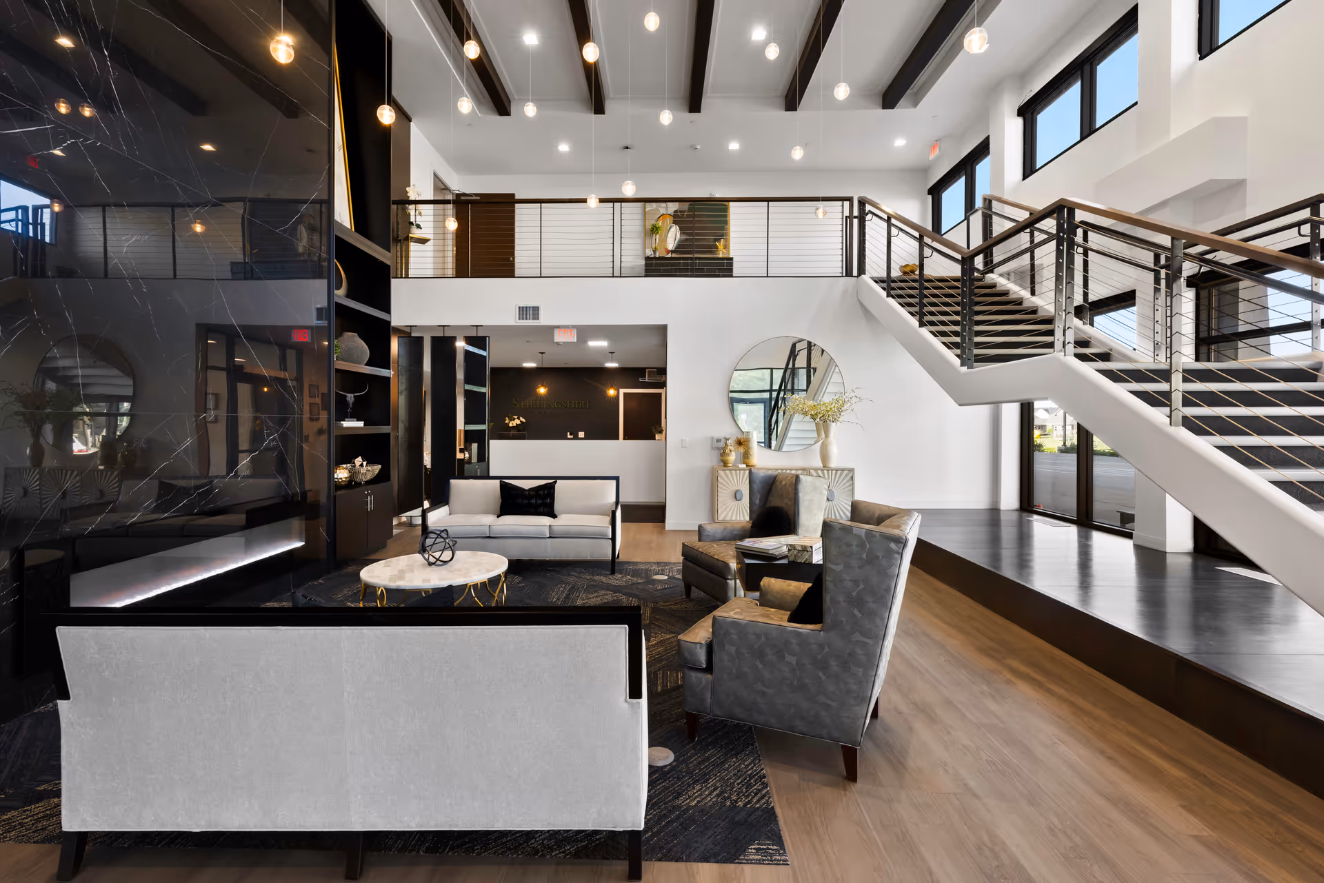 Modern senior living facility lobby with contemporary furniture including sofas and armchairs arranged around a round coffee table. The space features a high ceiling with hanging pendant lights, a staircase with metal railings leading to an upper level, large windows allowing natural light, and a reception desk in the background.