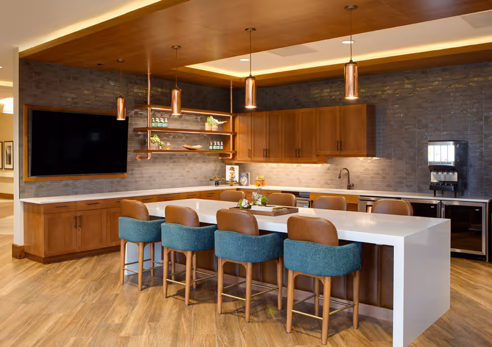 Modern kitchen area with a large white island countertop surrounded by six chairs, three with teal upholstery and three with brown leather. The kitchen features wooden cabinets, a dark tiled backsplash, a mounted flat-screen TV, pendant lights, and a beverage dispenser on the right side.