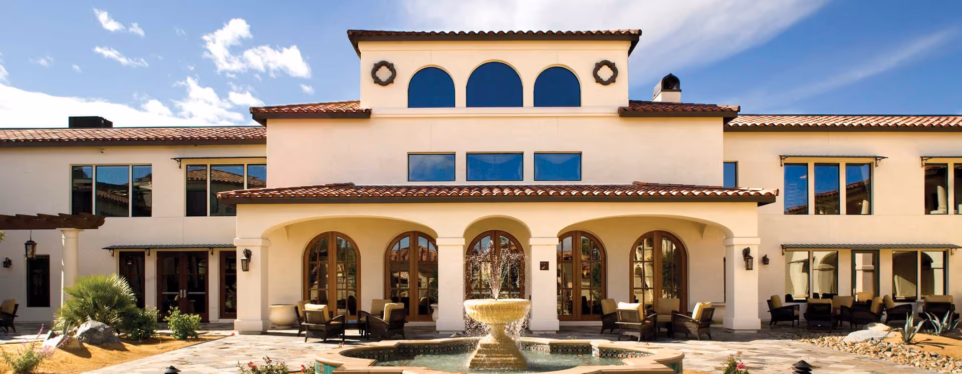 Exterior view of a two-story building with a tiled roof and arched windows and doorways. There is a fountain in the foreground surrounded by a paved area with outdoor seating and some landscaping including plants and rocks. The sky is clear with a few clouds.