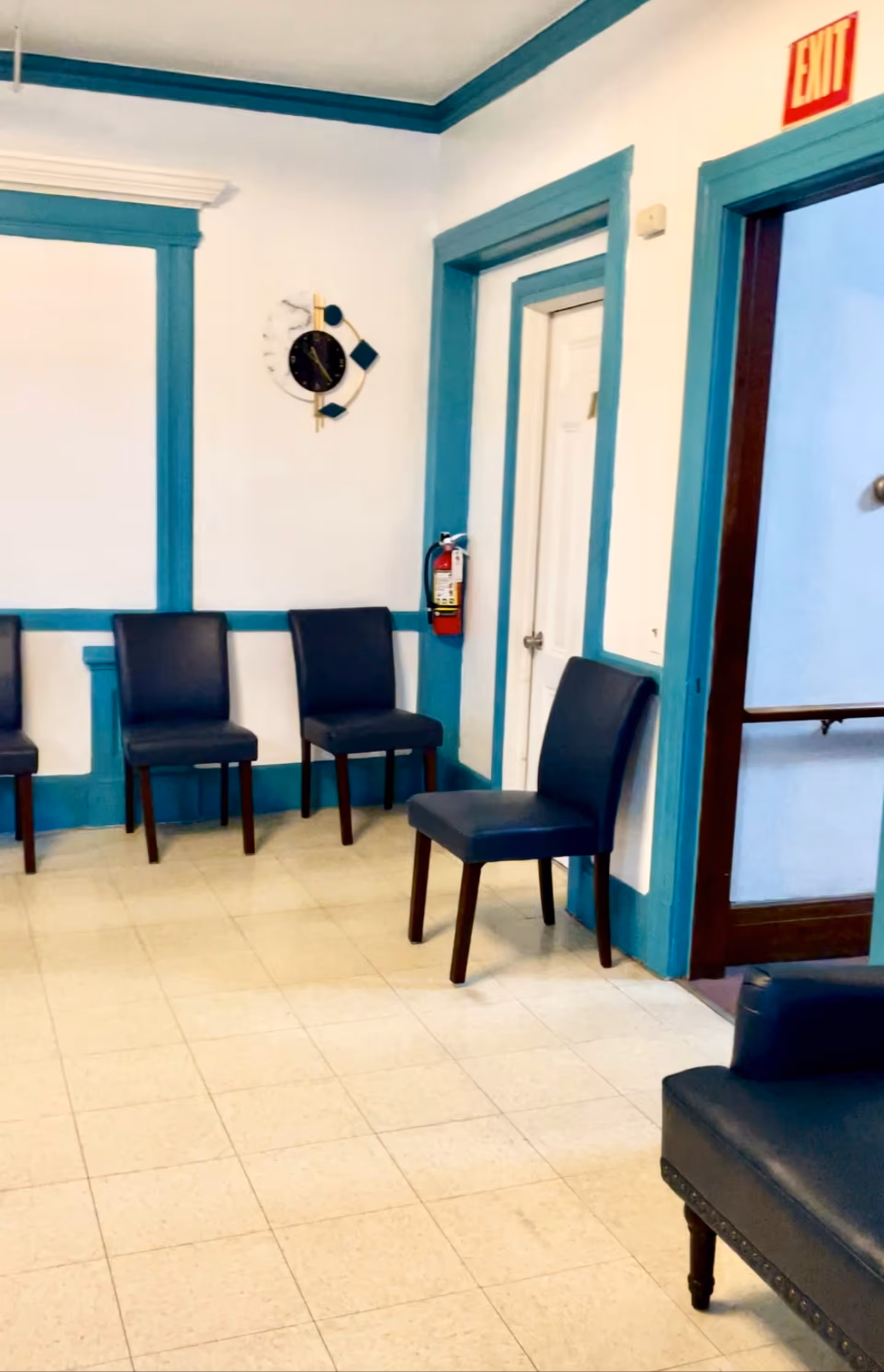 A small interior waiting area with blue-trimmed white walls, several dark blue chairs along the wall, a wall clock, a fire extinguisher, and an exit door.