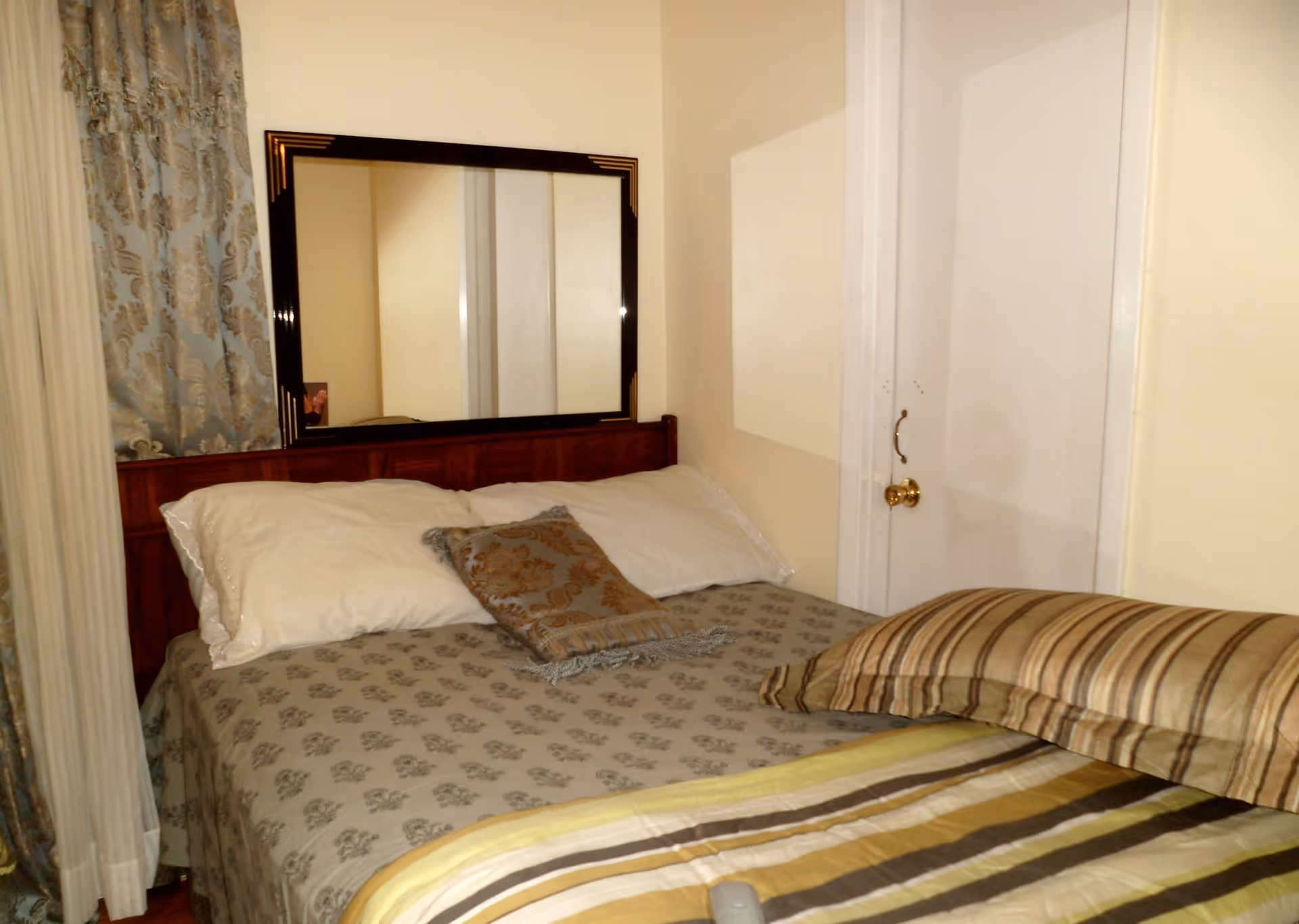 A bedroom with a bed covered in patterned bedspreads and pillows. There is a large mirror mounted on the wall above the headboard. Curtains with a floral design are visible on the left side, and a white door with a brass doorknob is on the right.