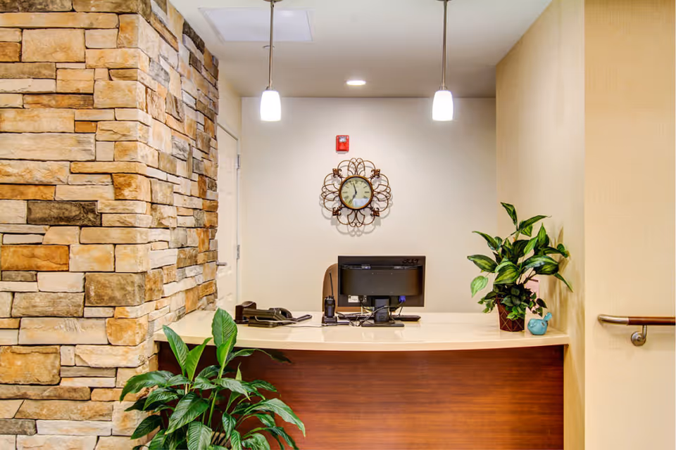 Reception desk area with a computer monitor, telephone, and two potted plants. The wall behind the desk has a decorative clock and a fire alarm. The left side features a stone wall, and two pendant lights hang from the ceiling above the desk.