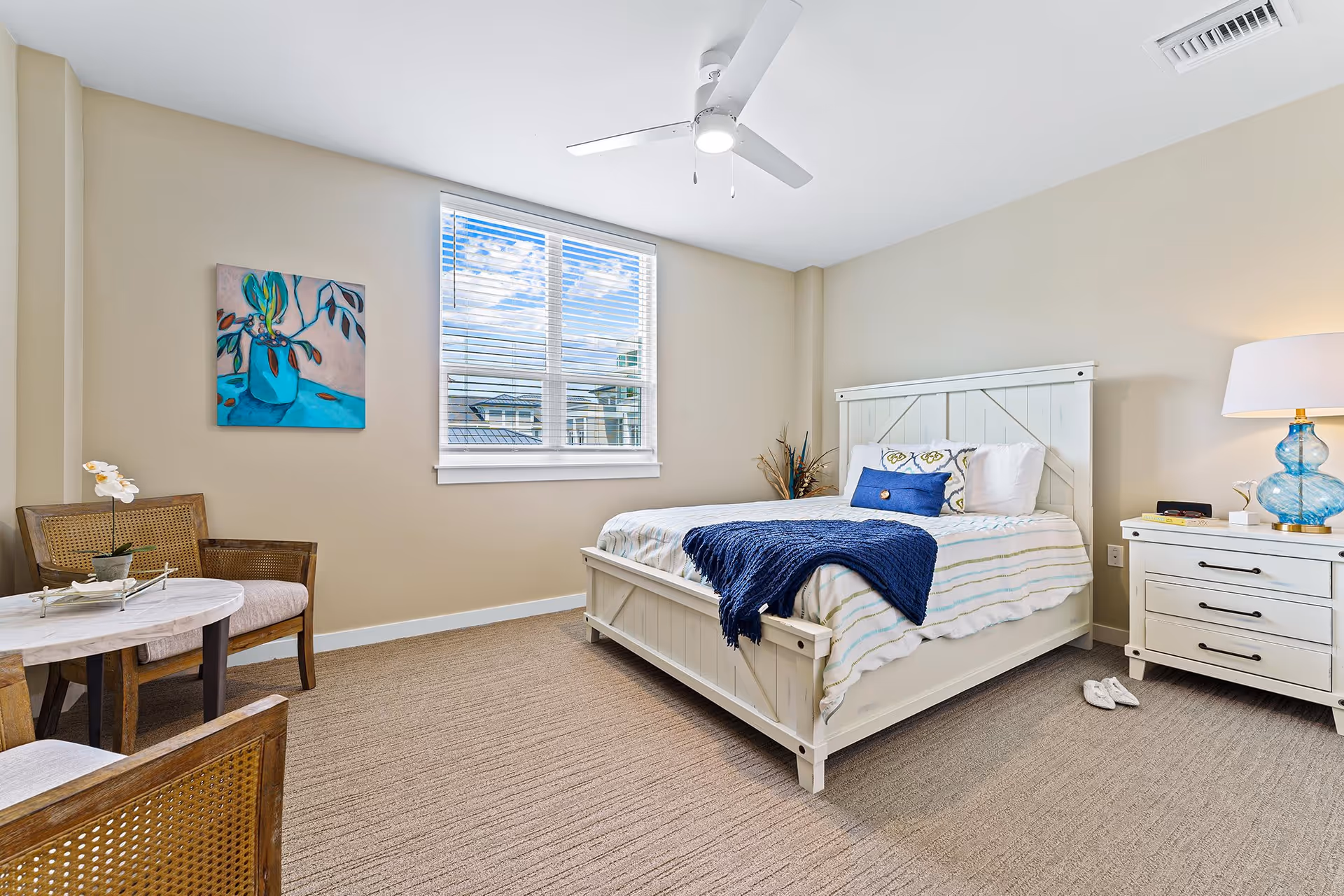 A bright and cozy bedroom with a white wooden bed frame, neatly made bed with striped bedding and a blue throw blanket. There is a white nightstand with a blue glass lamp and a pair of slippers on the carpeted floor. Two wooden chairs with cushions and a small round table with a potted orchid are placed near a window with white blinds. A colorful painting hangs on the beige wall.