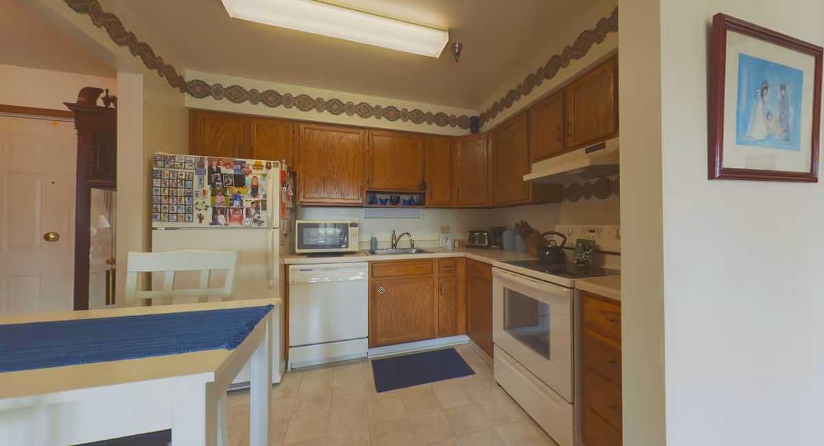 A kitchen with wooden cabinets, a white refrigerator covered with family photos and magnets, a microwave, dishwasher, stove with a kettle, and a sink. There is a white table with a blue runner and a white chair in the foreground. The walls have a decorative border near the ceiling, and a framed picture hangs on the right wall.