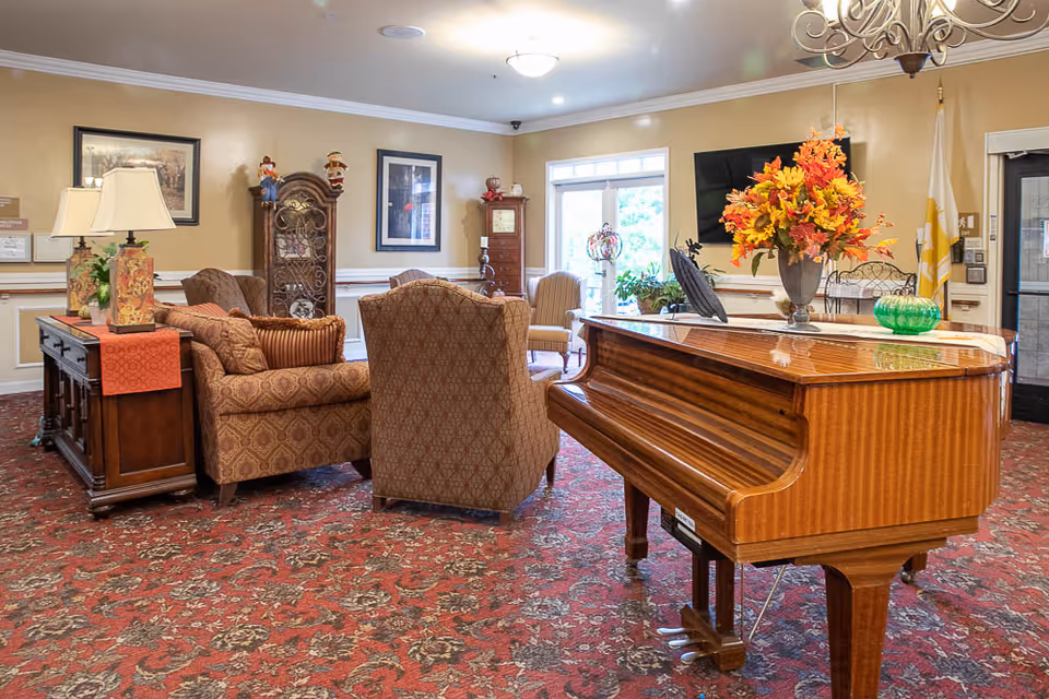 A cozy living room area in a senior living facility featuring a polished wooden grand piano adorned with a vase of colorful autumn flowers. The room has patterned red carpeting, upholstered armchairs and sofas, a wooden side table with two decorative lamps, framed artwork on the walls, and a large window letting in natural light.