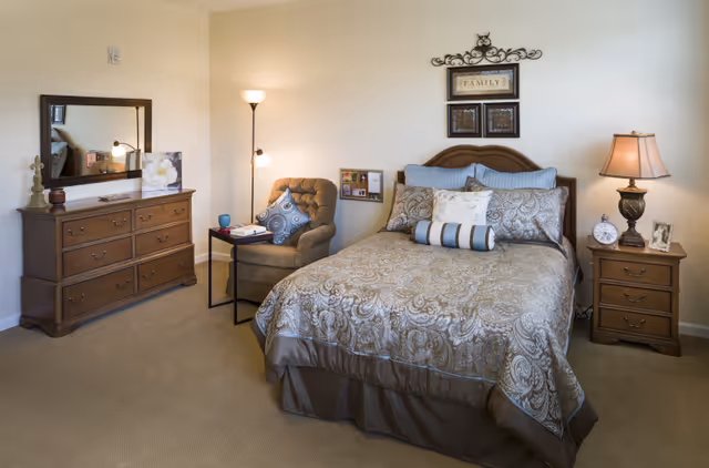 Neatly furnished bedroom with a bed, nightstand, dresser, upholstered chair, lamps, and wall decorations.