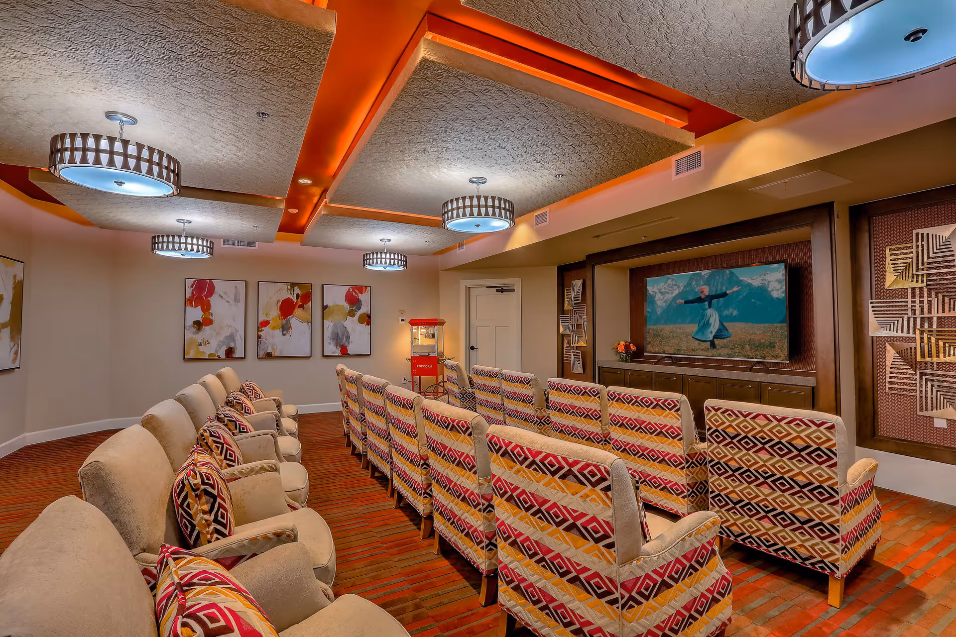 A cozy media room with two rows of cushioned chairs facing a large flat-screen TV mounted on the wall. The chairs have colorful patterned upholstery and matching pillows. The room features warm lighting with modern ceiling fixtures, abstract artwork on the walls, and a red popcorn machine in the corner.