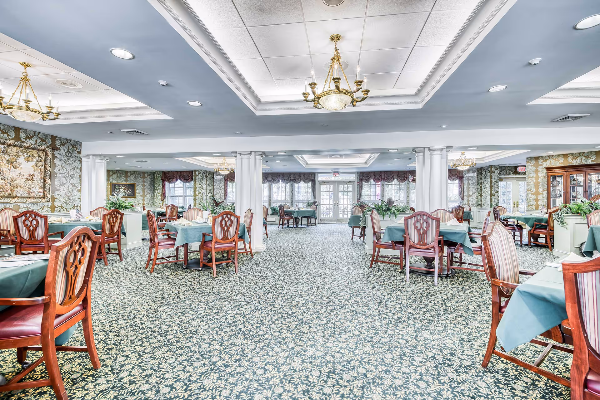 Spacious formal dining room with multiple tables and chairs, patterned carpet, chandeliers, and large windows.